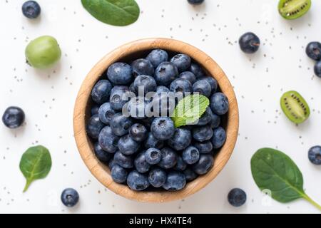 Heidelbeeren in Holzschale, Ansicht von oben. Flach zu legen oder Lebensmittel-Muster mit Heidelbeeren, Baby-Spinat, Baby Kiwi und Chia Samen auf weißem Hintergrund Stockfoto