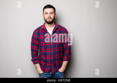 Hübscher junger Mann auf grauem Hintergrund Blick in die Kamera. Porträt des Lachens jungen Mannes mit Händen in den Taschen, graue Wand gelehnt. Glücklicher Kerl smi Stockfoto