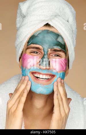 Glücklicher Mensch drei verschiedene Gesichtsmasken auf Gesicht auftragen. Foto des Mannes mit Handtuch auf dem Kopf auf beigen Hintergrund. Jugend und Skin Care Concept Stockfoto