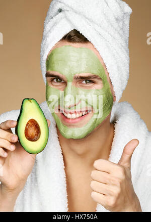 Porträt des jungen Mann mit Gesichtsmaske holding Slice von Avocado und Daumen auftauchen. Glücklich lächelnden Mann Spa-Behandlungen erhalten. & Schönheitshaut Auto Stockfoto