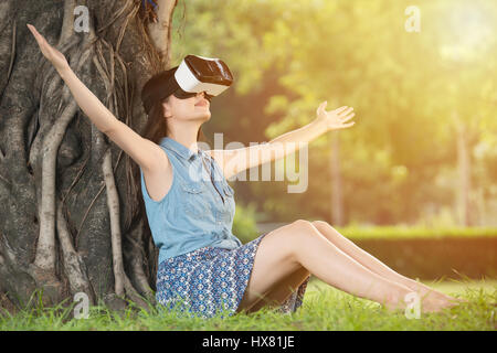 schöne asiatische Frau genießen Sie virtuell auf dem Rasen im Park im Freien sitzen. VR Kopfhörer Brille Gerät. Natur Natur Hintergrund. Menschen und techno Stockfoto