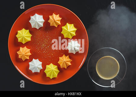 Mehrfarbig gemustert Cookies auf eine Runde orange Teller und eine Tasse heißen Kaffee auf einem schwarzen Hintergrund Stockfoto