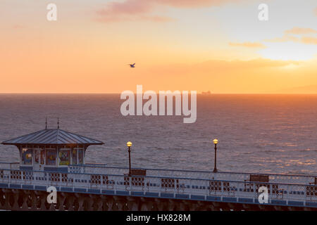 Ein goldener Sonnenaufgang wie es über dem Horizont mit einem Schiff in der Ferne und Llandudno Pier im Vordergrund bei dem Küstenort Llandudno bricht Stockfoto