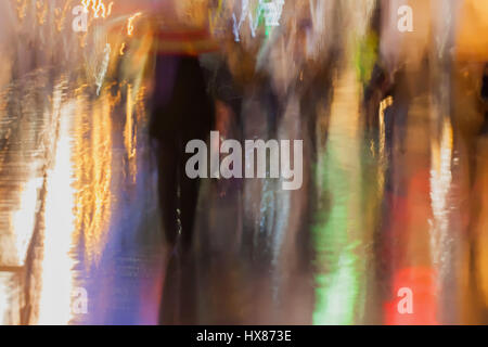 Abstrakter Hintergrund unscharf Menschen eilen nach unten die Stadtstraße unter Sonnenschirmen in regnerischen Abend, Impressionismus Stil, bunte Beleuchtung. Vorsätzliche Bewegungsunschärfe. Stockfoto