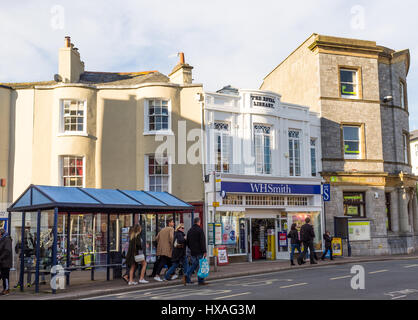 Eine Schlange von Menschen an einer Bushaltestelle direkt vor einem WH Smith-Shop in Teignmouth, Devon, UK. Stockfoto