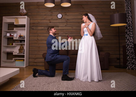 Bräutigam Anzug hält Trauringe, stehend auf die Knie und macht einen Vorschlag der Ehe, glückliche Braut im weißen Kleid und Schleier Stockfoto