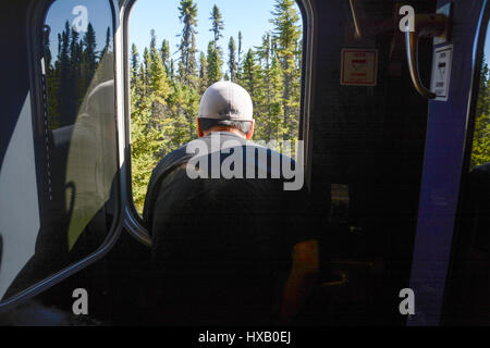 Ein indigener Cree-Mann blickt aus dem Fenster eines fahrenden Zugvorraums im borealen Wald in der Nähe der Stadt Moosonee, Ontario, Kanada. Stockfoto