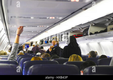 Ein Blick von der Rückseite der ein Passagierflugzeug der Kabine mit Menschen einsteigen, sitzen & Gepäckfächer verwenden, während Flugbegleiter schauen auf Stockfoto