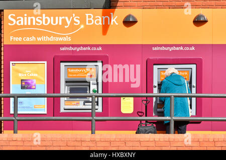 Zurück Ansicht graue Haare Reife Frau stehend auf Sainsburys Bank atm Loch in Wand Geldautomat Sainsburys Stadtzentrum Supermarkt Filiale Essex England UK Stockfoto