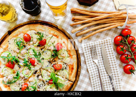 Leckere Pizza mit Käse, Tomaten und Fleisch, mit Glas Bier und dünne Baguette auf bedienten Tisch mit karierten Tischdecke Stockfoto