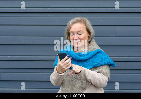 Attraktive Tech versierte Seniorin mit einer Handy-SMS eine Nachricht an einen Freund oder suchen eine Reihe zu nennen, grau Metall Hintergrund mit co Stockfoto