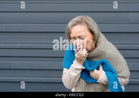 Elegante senior Frau Husten in die Hand und umklammerte ihre Brust in einem Konzept von saisonalen Bronchitis oder Grippe und medizinischen Gesundheitsversorgung, grau mich Stockfoto