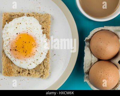 Flach Zusammensetzung und engen Ernte von einem pochierten Ei auf Toast, Sunny Side Up, Frühstück mit einer Tasse Tee oder Kaffee und Zwei rohe Eier in der Schale Stockfoto