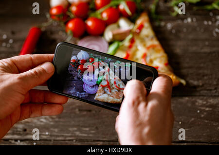Mann mit Smartphone zum Stück Pizza auf Holztisch fotografieren Stockfoto