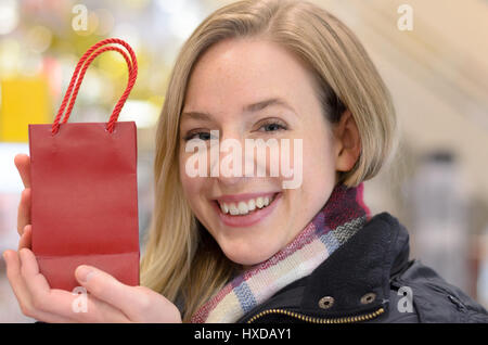 Glückliche Frau anzeigen ein kleines Weihnachtsgeschenk in eine festliche rote Shop-Tragetasche mit einem warmen Lächeln, Kopf und Schultern hautnah Stockfoto