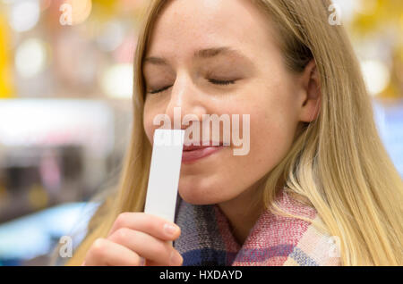 Junge, blonde Frau genoss ein Aroma mit einem schönen Lächeln der Glückseligkeit und Augen vor Lust geschlossen, da sie an eine weiße Karte in der Hand schnuppert Stockfoto