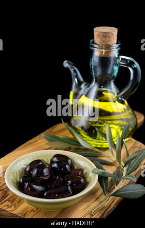 Schwarze Oliven, Flasche natives Olivenöl und Ölzweig auf Schneidbrett aus Holz und schwarzem Hintergrund Stockfoto