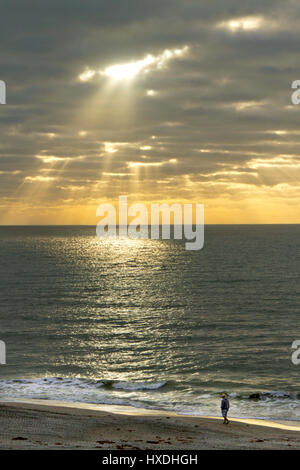 Lichtstrahlen unverdünnt in durch dunkle Wolken auf einem ruhigen Meer Iluminating einen leuchtenden Kreis des Lichts zu brechen, wie ein einsamer Mann am Strand geht Stockfoto