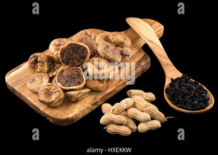Getrocknete Feigen auf Schneidbrett aus Holz, Erdnüsse und schwarze Rosinen in Holzlöffel auf schwarzem Hintergrund Stockfoto