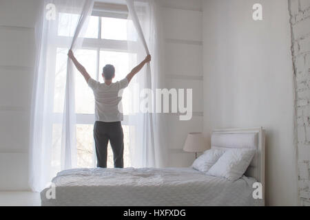 Junger Mann Eröffnung Vorhänge zu Welcomw Morgen und Licht, rückseitige Ansicht Stockfoto