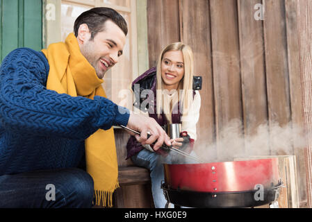 Lächelnde junge Frau mit Heißgetränk betrachten glücklicher Mann Grillen von Fleisch auf Veranda Stockfoto