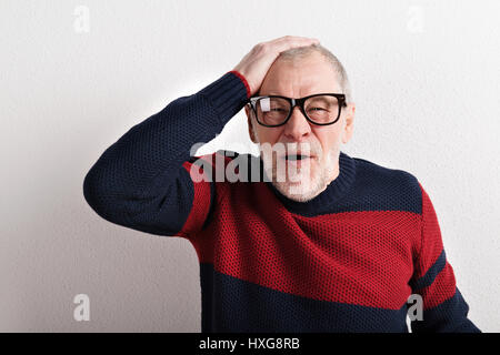 Aufgeregt senior Mann mit Kopfschmerzen Kopf haltend, roten und blauen Pullover und schwarze Brillen tragen. Studio gegen weiße Wand erschossen. Stockfoto