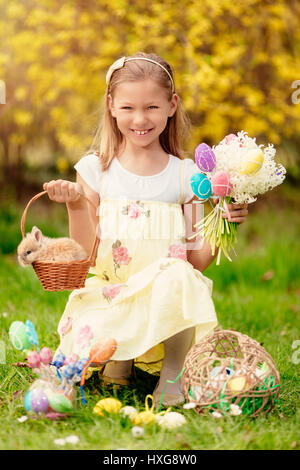 Schönen lächelnden kleinen Mädchen Holding niedlichen Häschen in den Korb und Strauß weißer Blumen und Ostereier in den Frühlingsferien. Blick in die Kamera. Stockfoto