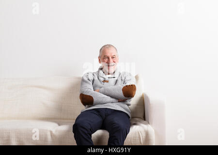 Gut aussehend senior lächelnder Mann in grauen Pullover, überquerte sitzt auf dem Sofa, ruhen, Arme. Studio gegen weiße Wand erschossen. Stockfoto