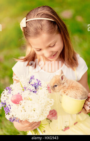 Wunderschönes lächelndes Mädchen mit niedlichen Häschen und Strauß weißer Blumen und Ostereier in Frühlingsferien. Stockfoto