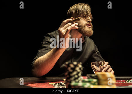 Bärtiger Mann mit Zigarre und Glas am Pokertisch sitzen und wegsehen Stockfoto