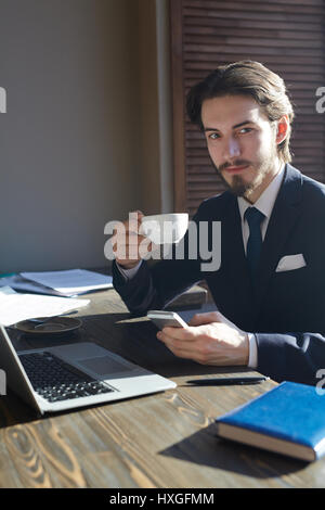 Porträt der schönen eleganten Geschäftsmann arbeiten mit Smartphone und Laptop an sonnenbeschienenen Holztisch während der Kaffeepause, Blick in die Kamera und lächelnd Stockfoto