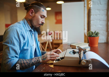 Seite Ansicht Porträt des stilvollen tätowierter Mann mit Oldschool Vintage Schreibmaschine auf Holztisch im Café schreiben Geschichte Stockfoto