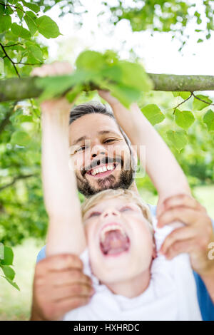 Vater Sohn Aufstieg Baum hilft und sie Spaß haben, Lachen im Sommer Stockfoto