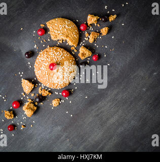 Haferflocken-Cookies sind ihre Stücke und Krümel mit roten Beeren auf einem schwarzen Hintergrund aus Holz. Platz für den Text. Zurückhaltend. Stockfoto