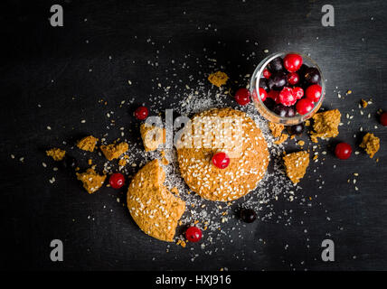 Haferflocken-Cookies sind ihre Stücke und Krümel mit roten Beeren auf einem schwarzen Hintergrund aus Holz. Platz für den Text. Zurückhaltend. Stockfoto