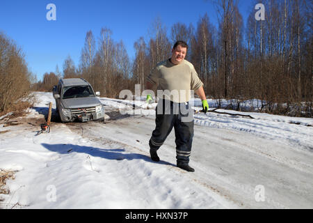 Nazia Village, Leningrad Region, Russland - 17. März 2015: Auto stecken auf einem Waldweg, Vorderrad verfiel Schlucht im Eis, der Mann zieht das Kabel Stockfoto