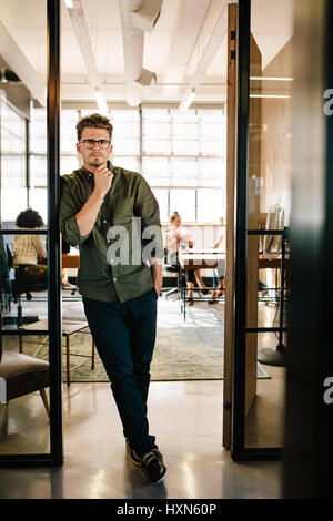 In voller Länge Portrait von gut aussehenden jungen Mann stehend in Tür des Büros mit Menschen, die im Hintergrund arbeiten. Stockfoto