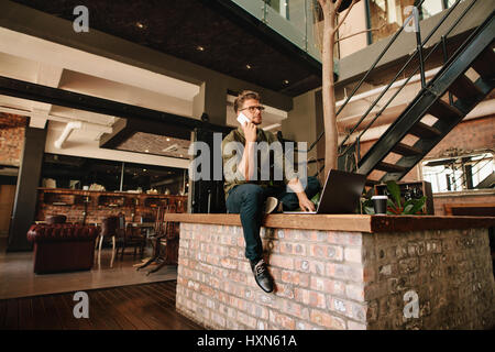 Kreative junge Mann sitzt im Büro Cafeteria mit Laptop und telefonieren mit Handy. Stockfoto