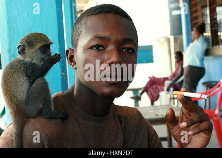 Zanzibar, Tansania - 16. Februar 2008: Junge afrikanische Mann 25 Jahre alt, in einem Straßencafé, Rauchen eine Zigarette zu halten, zähmen, Baby sittin Green monkey Stockfoto