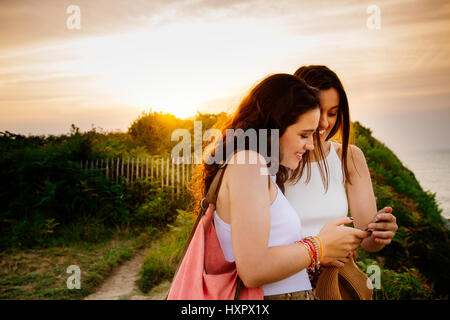 Glückliche junge Freundinnen beobachten etwas in das Smartphone auf einer Klippe bei Sonnenuntergang Stockfoto