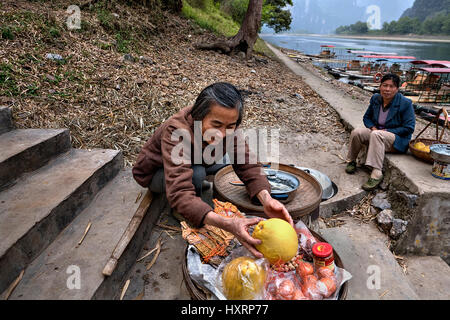 Yangshuo, Guangxi, China - 29. März 2010: Lächelnde ältere Asiatin Landwirt verkauft Obst auf Stufen zum Pier auf Hintergrund von Bambus Flöße und Stockfoto