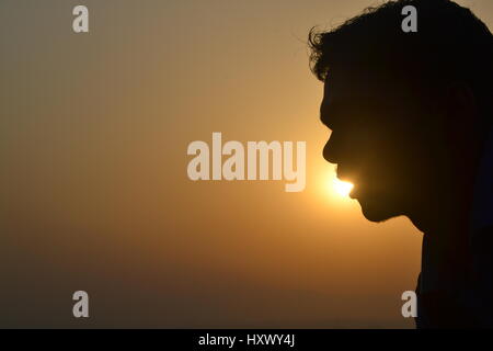 Seitenprofil-Silhouette eines jungen Indianers bei Sonnenuntergang mit der Sonne in seinem offenen Mund, wodurch eine kreative, perspektivische Beleuchtung entsteht Stockfoto