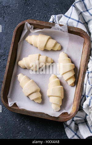 Rohe ungebackenen Croissant auf Backpapier in alten Backblech mit Küchentuch über schwarze Textur konkreten Hintergrund. Ansicht von oben Stockfoto
