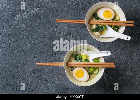 Zwei Schüsseln mit asiatischen Stil Suppe mit Rührei, die Hälfte der marinierte Ei, Frühlingszwiebeln, Spinat serviert mit hölzernen Stäbchen und Löffel über schwarze te Stockfoto