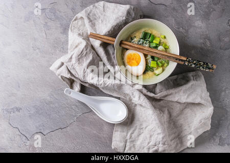 Weiße Schale mit asiatischen Stil Suppe mit Rührei, die Hälfte der marinierte Ei, Frühlingszwiebeln, Spinat serviert mit hölzernen Stäbchen, Löffel, Textile über gra Stockfoto