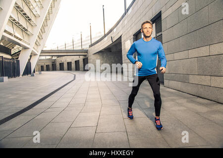 Hübscher Mann mittlerer Alter ernst in blau schwarz Sport-Uniform und Kopfhörer und Fitness Tracker läuft in der Stadt. Stockfoto