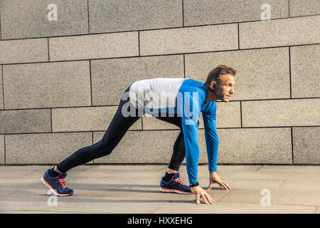 Hübscher Mann mittlerer Alter ernst in blau schwarz Sport-Uniform und Kopfhörer und Fitness Tracker läuft in der Stadt. Stockfoto