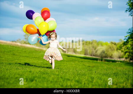 Kleine blonde Mädchen laufen mit Haufen von bunten Luftballons auf dem Lande Stockfoto