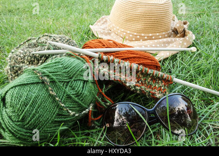 Außenansicht des Strickens Elemente wie stricken Garn und Nadeln, Sonnenbrille und Sommerhut auf Rasen als konzeptuelle Ansicht von Hobby und Freizeit Stockfoto