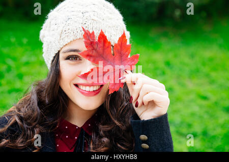 Schöne junge Frau mit Wollmütze für Gesicht mit einem roten Blatt während lächelnd vor einem grünen Hintergrund. Frische Haut und gesundes Lächeln. Stockfoto